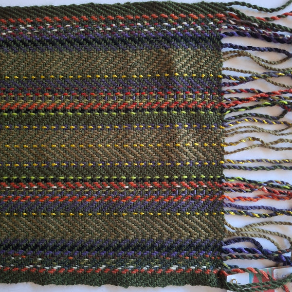 A close-up of a woven scarf featuring stripes in olive green, purple, rust red, white, and yellow, finished with fringed edges on the right.