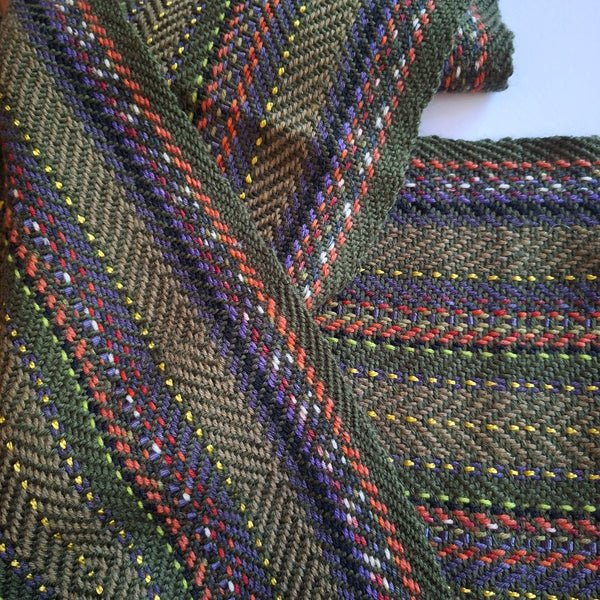 A close-up of a woven scarf with a pointed twill pattern in olive green, purple, and multicolored stripes of red, white, yellow, and purple.