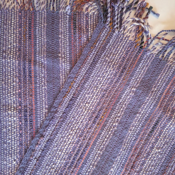 A close-up of a woven textile with fringe, showing a heathered mauve base with thin vertical stripes in muted  pink, and white.